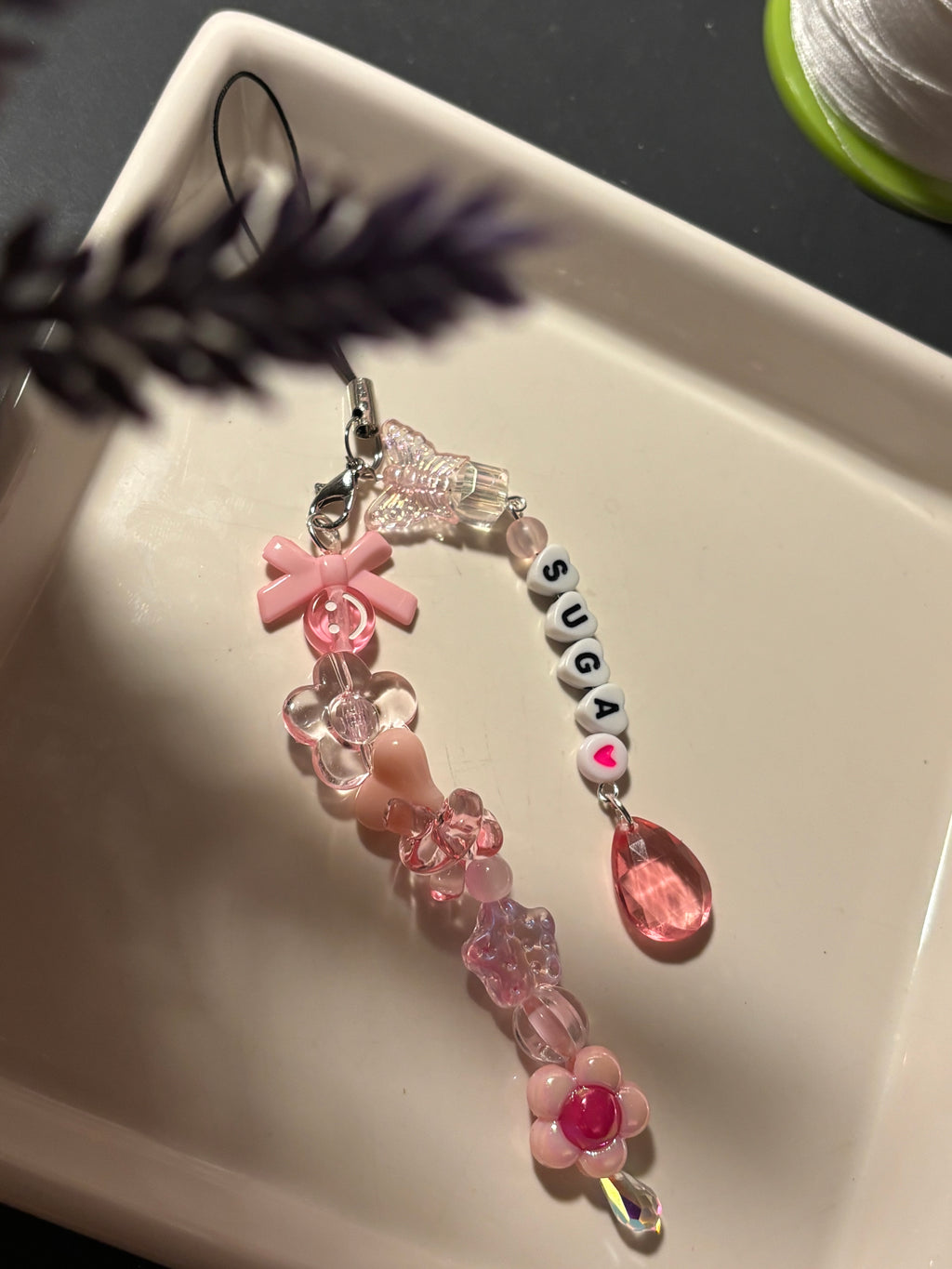 A pink beaded charm keychain with a butterfly charm, pink bow, clear flower beads, white letter beads spelling 'SUGA', and a pink teardrop charm, resting on a white ceramic tray.