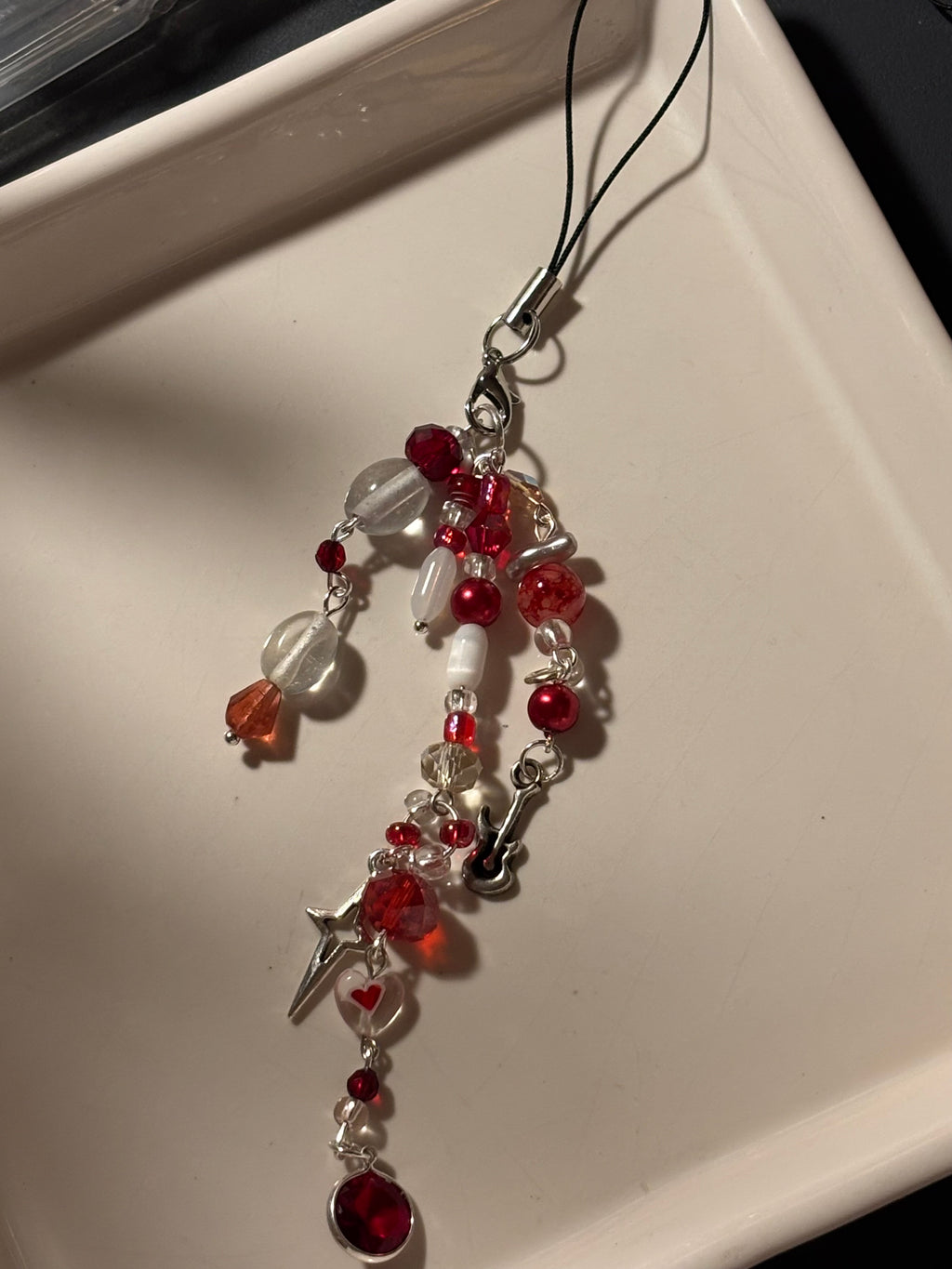 A beaded charm strap with red and clear beads, a silver clasp, and dangling charms including a small silver cross and heart details, resting on a white tray.
