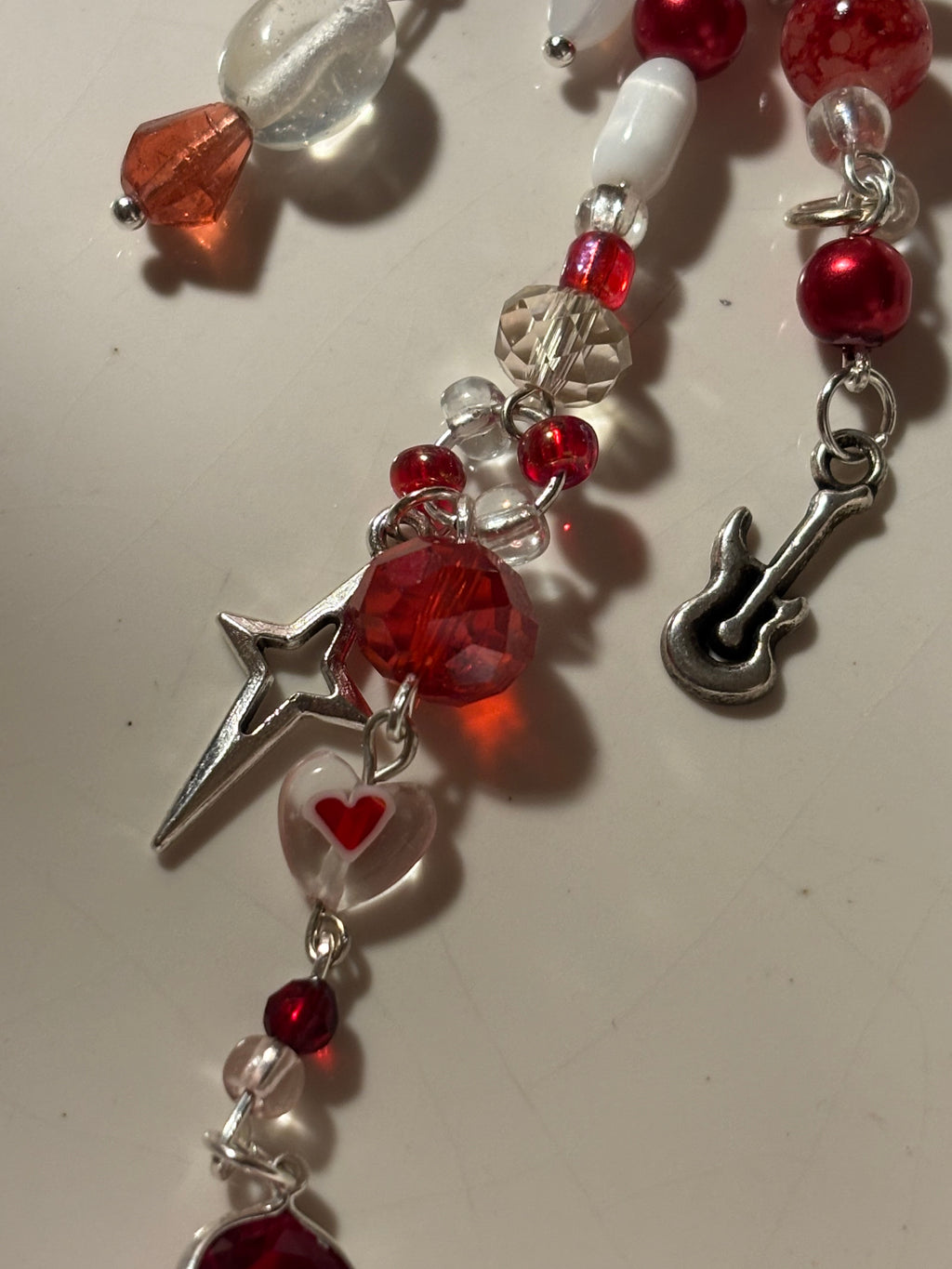 A close-up of a beaded jewelry strand featuring red faceted beads, clear glass beads, a small silver guitar charm, and a silver star charm on a light beige background.