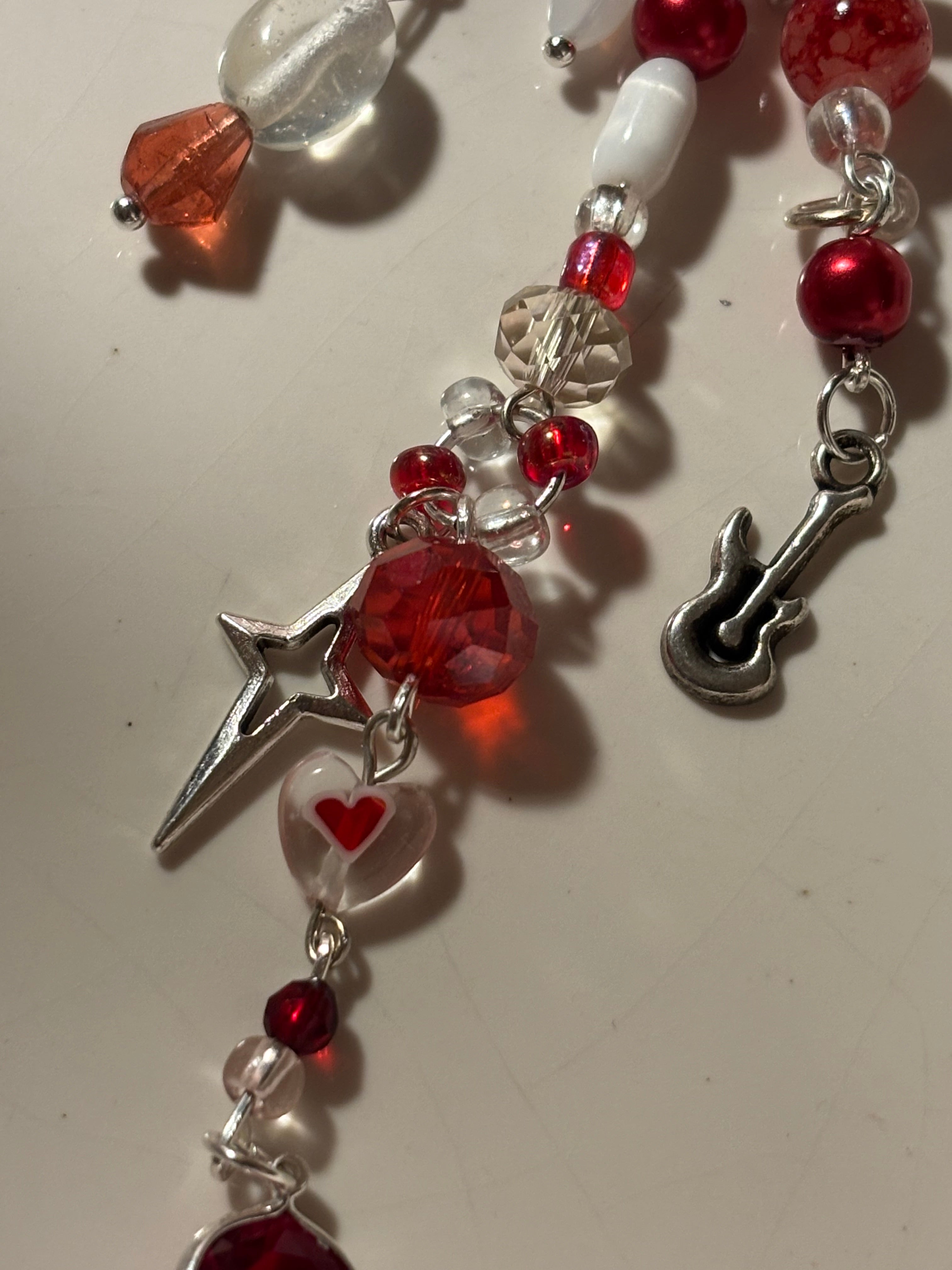 A close-up of a beaded jewelry strand featuring red faceted beads, clear glass beads, a small silver guitar charm, and a silver star charm on a light beige background.