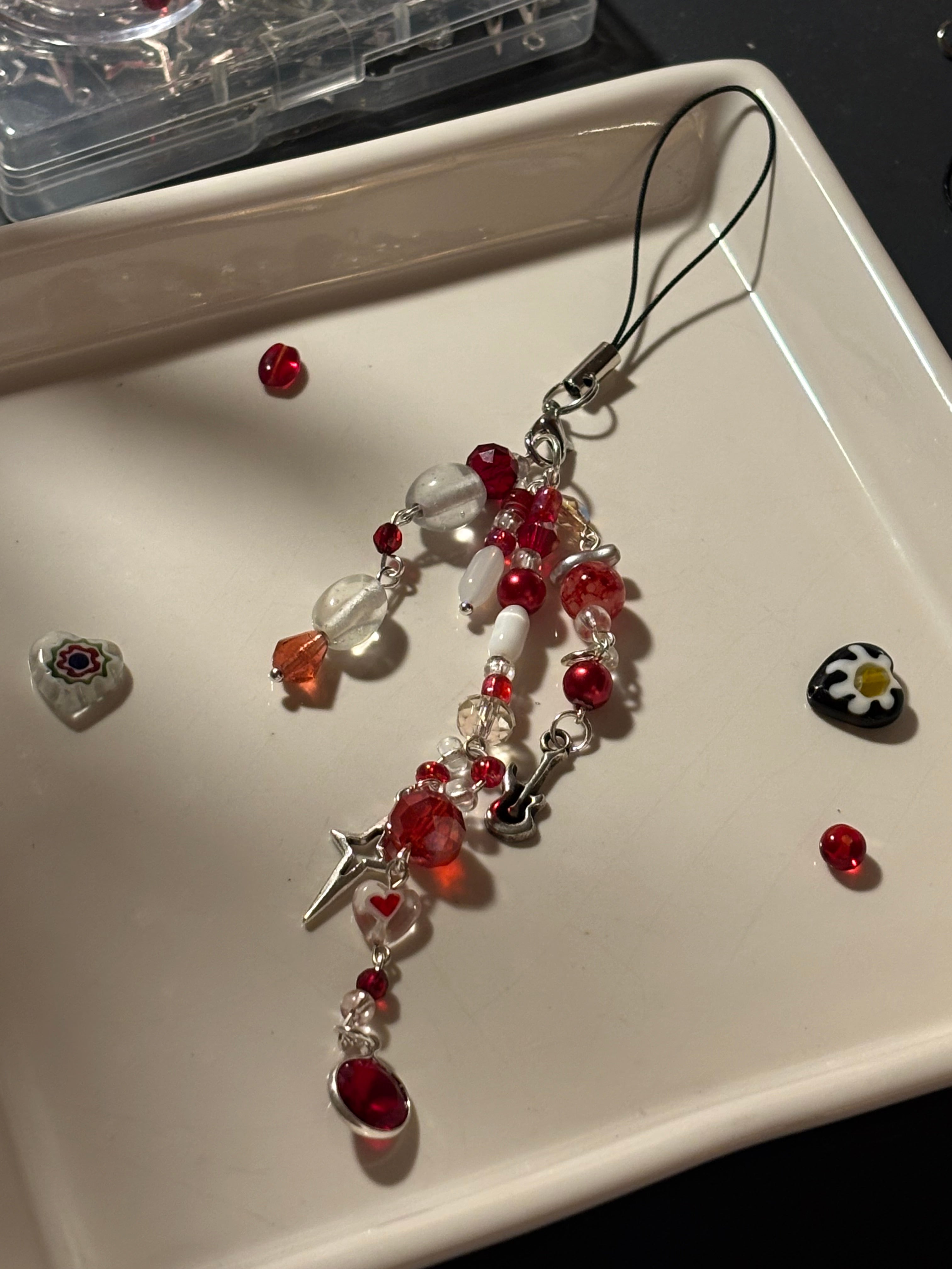 A handmade beaded charm or phone strap with red and clear beads, silver-toned metal, and small charms, displayed on a white tray.