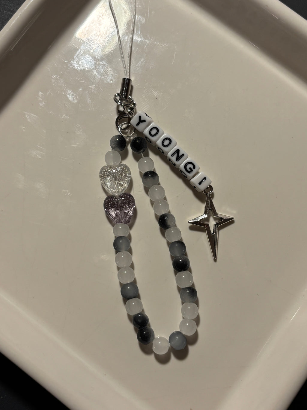 A personalized beaded phone strap or charm with the name 'YOONGI' in white letter beads, gray and white round beads, a clear heart charm, a lavender heart charm, and a silver star charm, displayed on a white ceramic dish.