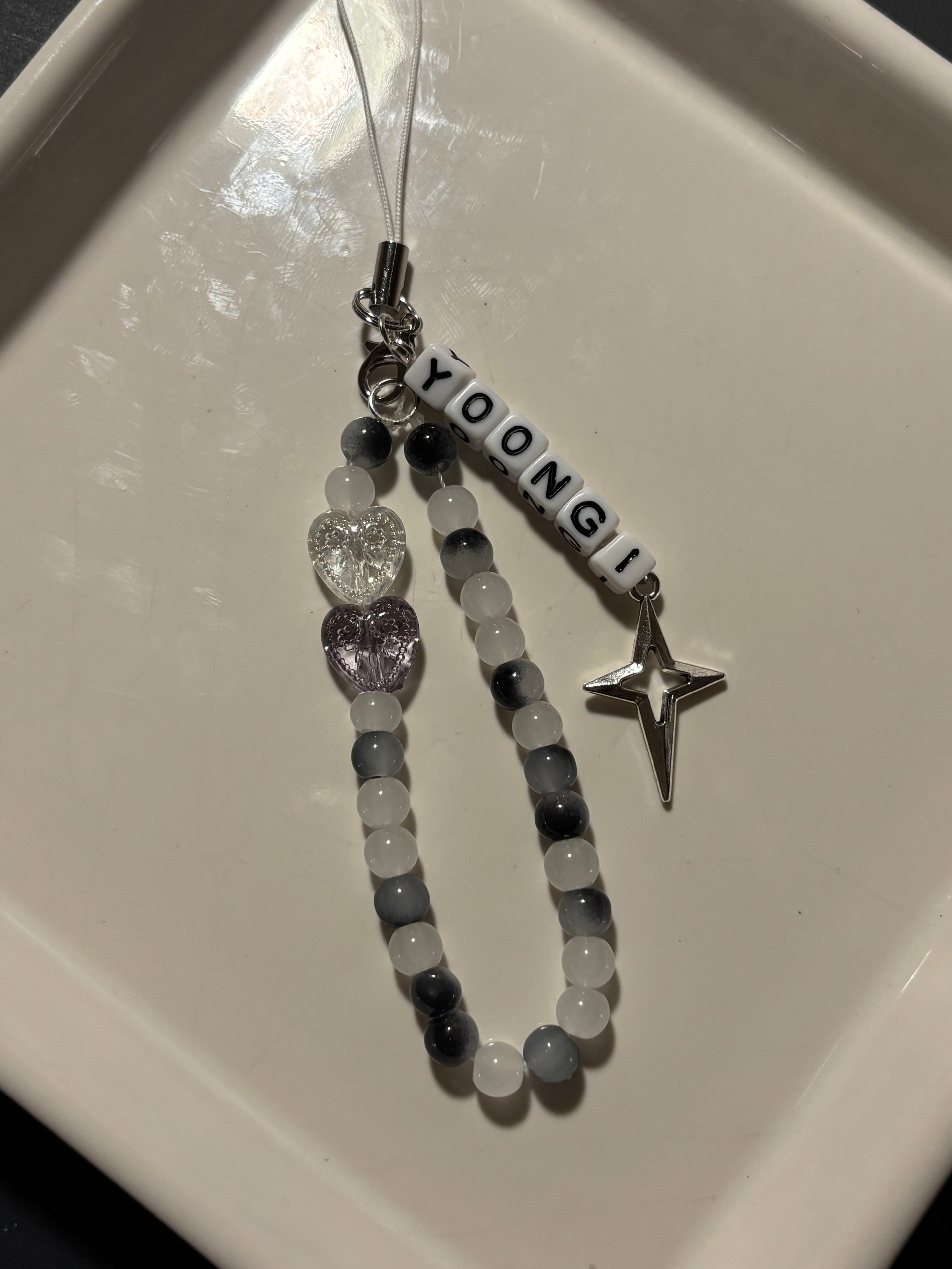 A personalized beaded phone strap or charm with the name 'YOONGI' in white letter beads, gray and white round beads, a clear heart charm, a lavender heart charm, and a silver star charm, displayed on a white ceramic dish.