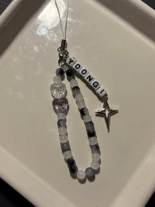 A personalized beaded phone strap or charm with the name 'YOONGI' in white letter beads, gray and white round beads, a clear heart charm, a lavender heart charm, and a silver star charm, displayed on a white ceramic dish.